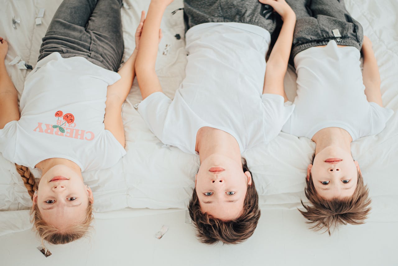 services-07 Children lying on a white bed, wearing casual white t-shirts. Bright and playful indoor setting.