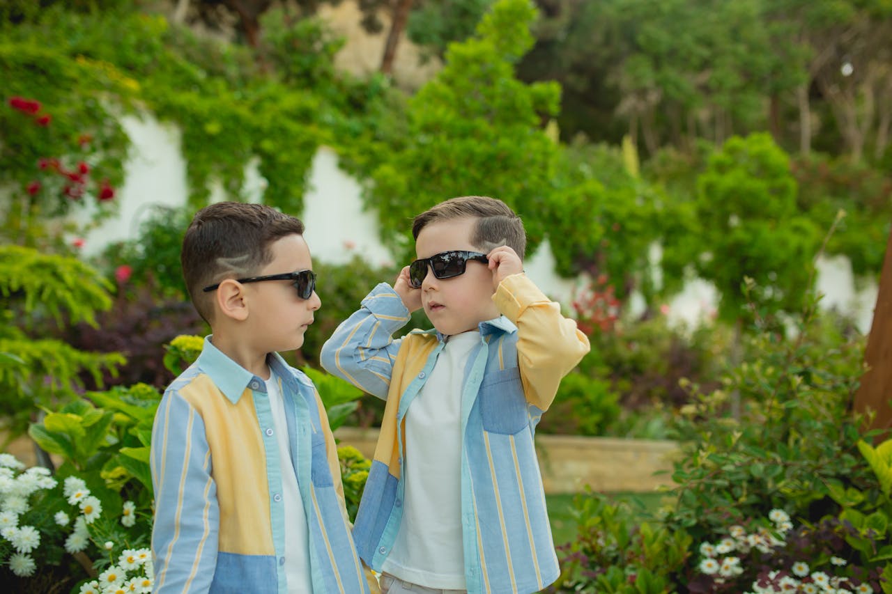 Two children wearing sunglasses and matching outfits enjoying a sunny day in a lush garden.