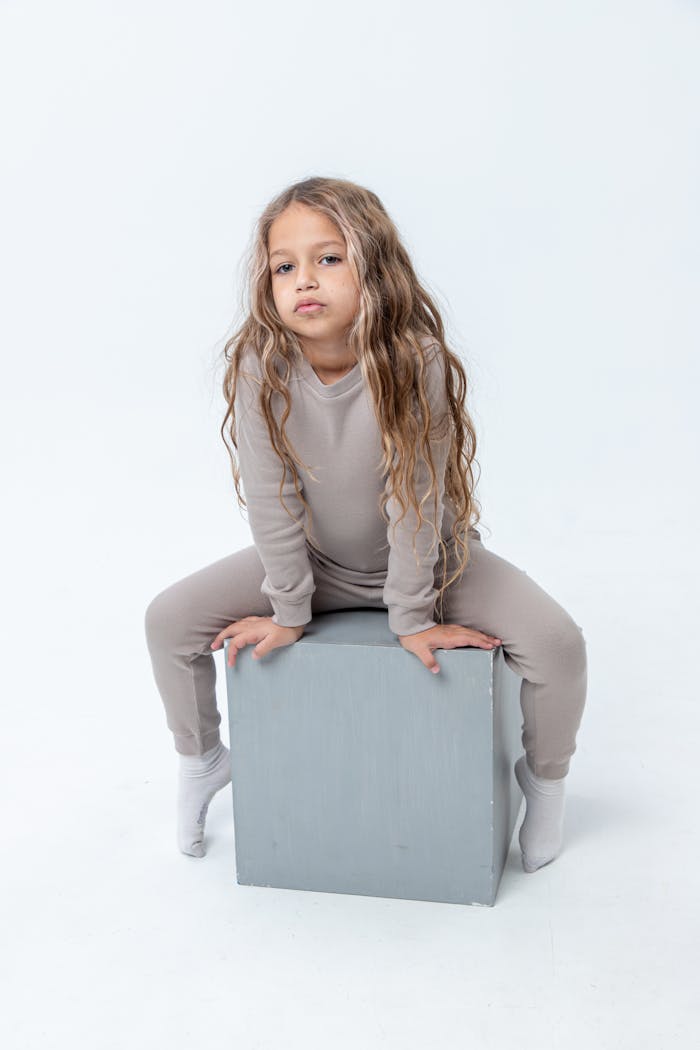 Child with long blonde hair sitting on a box in a studio with a white background.