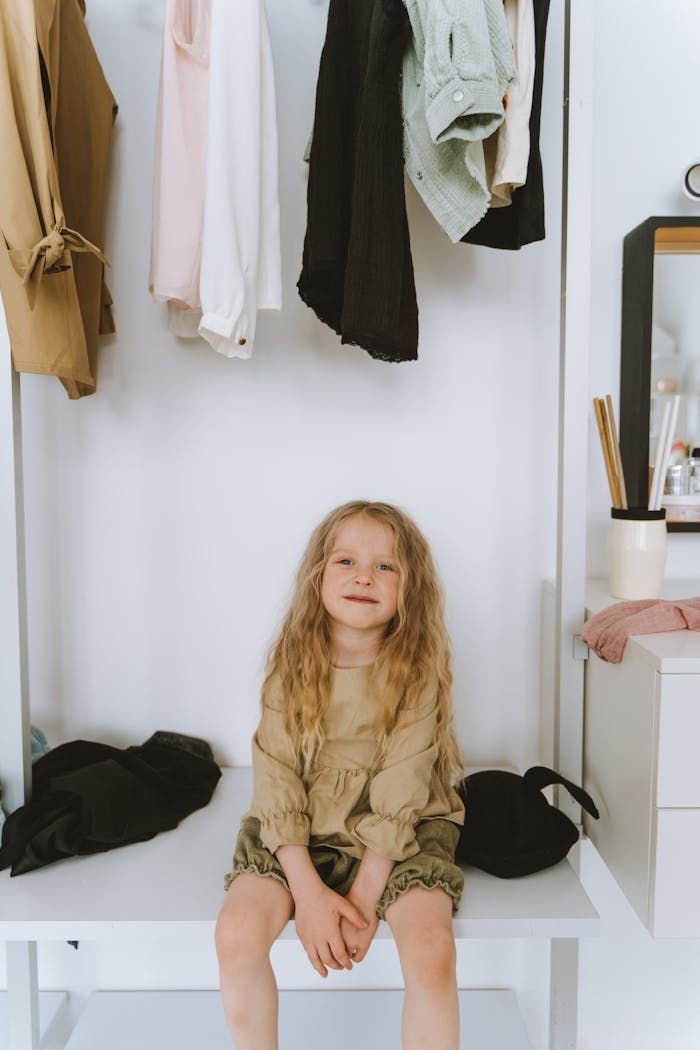 services-06 A cheerful young girl seated in a wardrobe area with clothes hanging around her indoors.